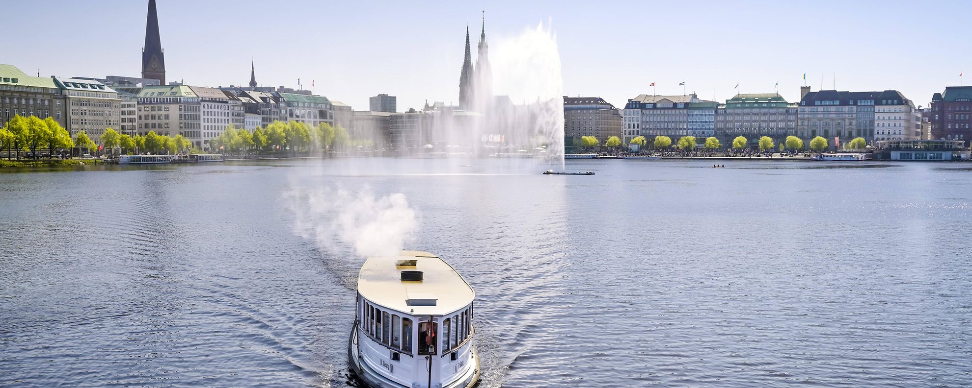 Auf der Binnenalster fährt ein Alsterdampfer. Weiter hinten sprudelt die Alsterfontäne in die Höhe.